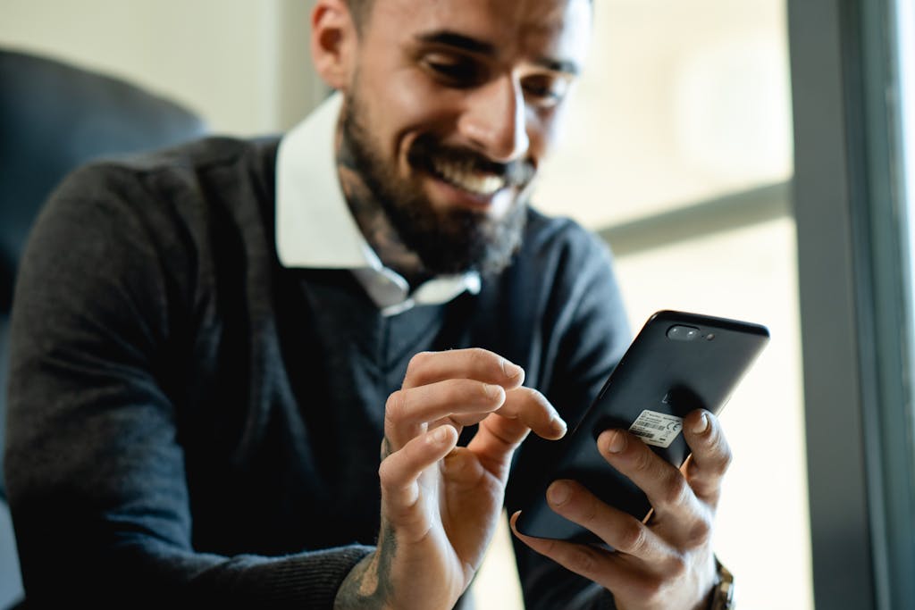 A bearded man smiles while using his smartphone inside a well-lit room.